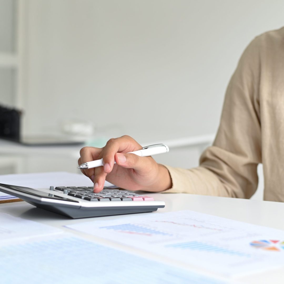 A young man with a pen in hand is operating a calculator, graphing data and laptop on the desk.